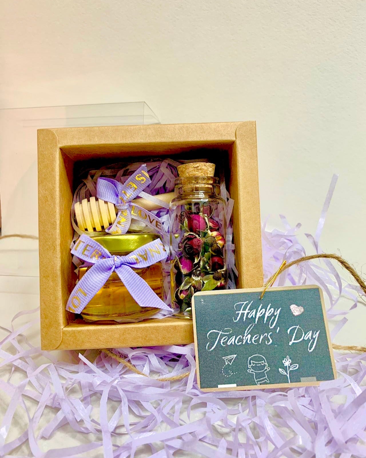 A gift set containing a jar of honey and a packet of rose tea, presented in a brown box with a purple ribbon and a 'Happy Teacher's Day' tag.