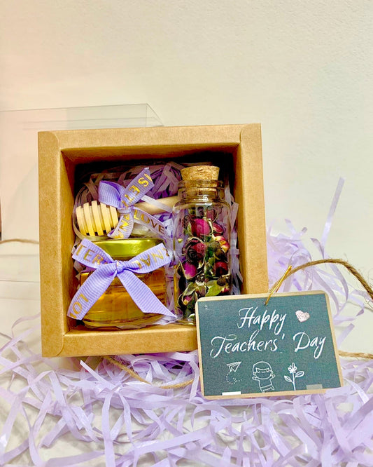 A gift set containing a jar of honey and a packet of rose tea, presented in a brown box with a purple ribbon and a 'Happy Teacher's Day' tag.